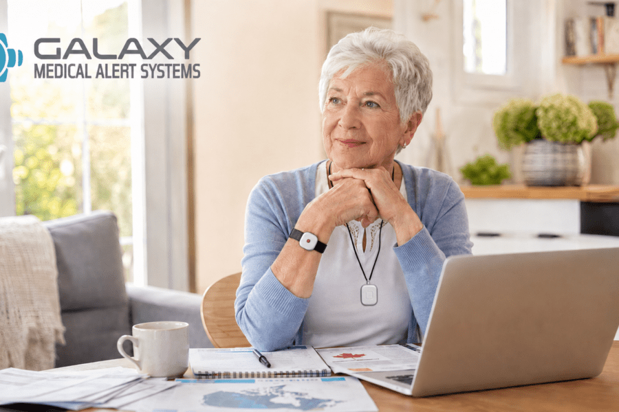 Senior in Canada wearing a Galaxy medical alert system pendant and wrist button, reviewing medical alert system coverage options on a laptop